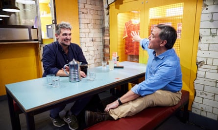 Two men, both wearing blue shirts, sitting at a restaurant table talking