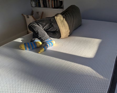 A man lies on his side with his back to the camera on a mattress and bed in a bedroom