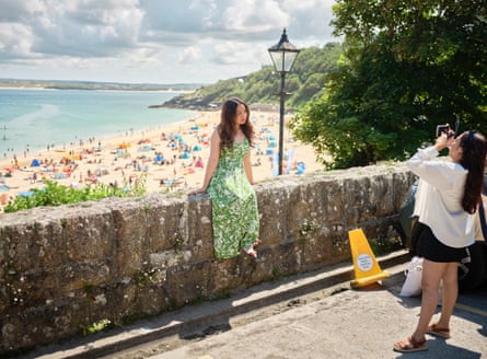 A woman taking a photograph of anpther woman sitting on a wall in St Ives, Cornwall, UK, with a crowded beach behind her, July 2024