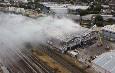Smoke billows from the roof of a large industrial unit by the side of railway tracks.