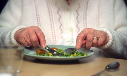 An elderly woman's hands hold a knife and fork over a plate of food