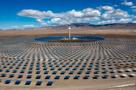A tall, tin white tower in a desert landscape is surrounded by thousands of mirrors and solar panels. There are small hills in the background and a deep blue sky with white fluffy clouds.