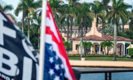 A view of the pink buildings of Donald Trump's Mar-a-Lago estate, surrounded by palm trees. In the foreground, an American flag can be seen.