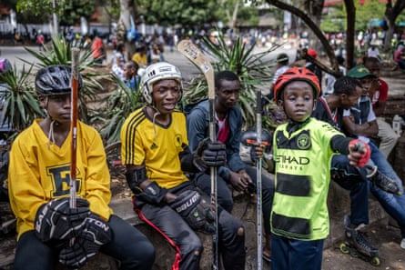 three boys, two in yellow jerseys, sit holding hockey sticks in a car park; they wear gloves and helmets.