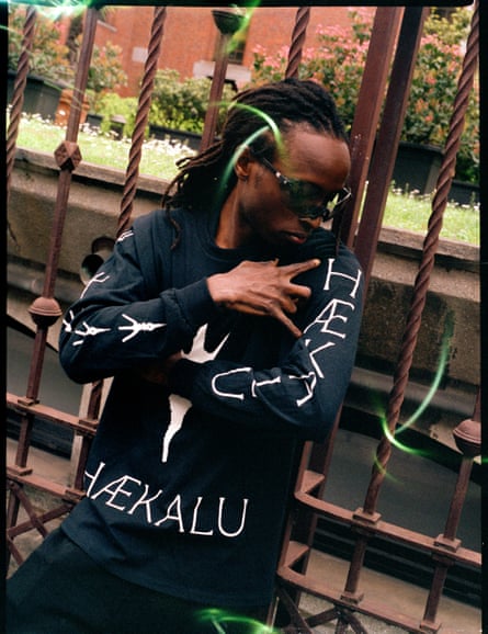 Martin Kanja leans back against a metal fence and looks down at the word ‘Haekalu’ on the sleeve of the black top he is wearing