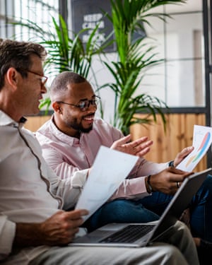 Two businessmen using laptop and discussing documents on sofa in loft office.