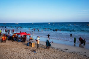 People standing on a beach, sitting on plastic chairs, or wading in the water