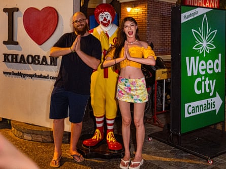 A man and a woman pose with their hands in prayer in imitation of a praying Ronald McDonald statue, next to a sign saying ‘Weed City’.