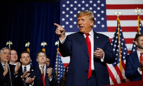 man wearing blue suit and red tie points finger as men in suits applaud behind him
