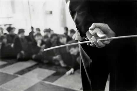 The hands of a teacher are seen from behind holding a cane in front of a class full of students.