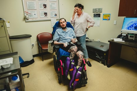 A woman holding her hands to her face beside her son in a wheelchair in a doctor’s office