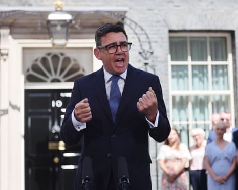 Andy Burnham in dark suit and blue tie stands at microphone with both fists tightened in front of him