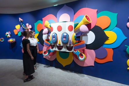Two women stand in front of a colourful semi-sculptural exhibit with a face surrounded by horns and petals