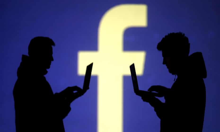 Silhouettes of laptop users are seen next to a screen projection of Facebook logo