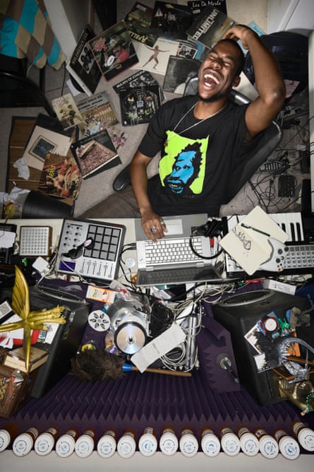 Flying Lotus, with a huge grin, seen from above with recording equipment on his desk and papers on the floor behind him