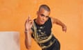 Layton Williams posing on one leg and leaning forwards, as if he is dancing, wearing a black vest embellished with gold, a gold earring, black trousers and boots, and photographed in an orange space with white marbled blocks