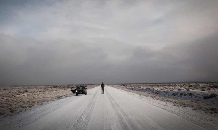 Parked car with lonely figure in the distance. Road is otherwise empty and landscape bleak and wintry.