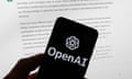 OpenAI logo is seen on a mobile phone in front of a computer screen displaying output from ChatGPT