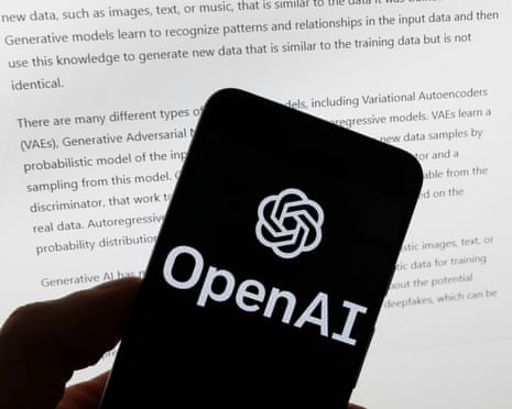OpenAI logo is seen on a mobile phone in front of a computer screen displaying output from ChatGPT