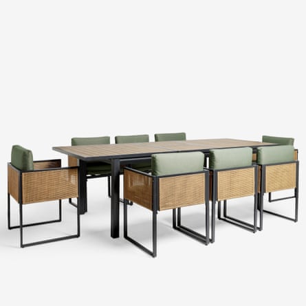A long wooden table with four legs is surrounded by 8 chairs that have black metal frames, rattan sides and backs and sage green cushions.