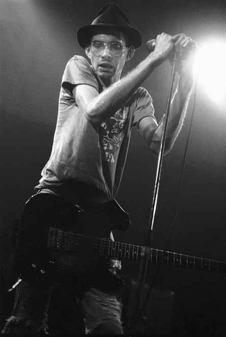 Steve Albini, with round glasses, a hat, T-shirt and guitar at his waist, sweats during a performance and clutches a microphone