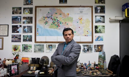 Israel Ticas in his office, standing in front of images of his team members at work and a map of El Salvador marking points of reported violent crimes.