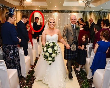 A bride and groom walk down the aisle hand-in-hand as the unknown guest looks on.