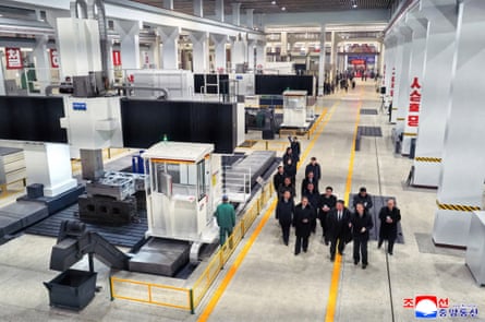 he walks in a group down an aisle between white pillars and what looks like machinery testing equipment.