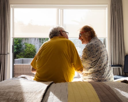 Back shot of a senior couple sitting on the edge of a bed, in front of a window.