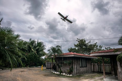 Uma aeronave leve voando baixo sobre uma casa de madeira de um andar cercada por palmeiras