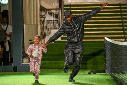 Skepta wearing a black jacket, matching trousers and cap runs across a stage with his young daughter who is wearing a pink snakeskin patterned matching top and trousers