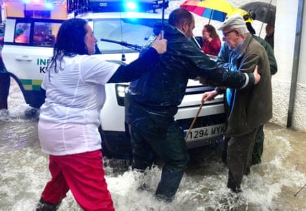 Water running in street, emergency vehicle and man with stick being helped by emergency worker with woman holding umbrella