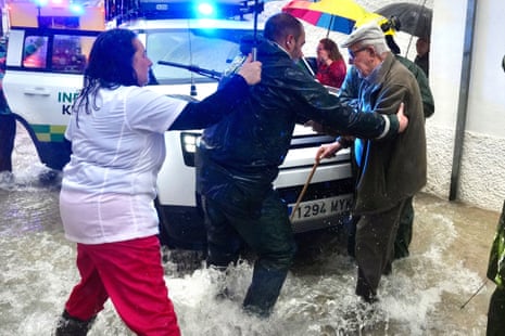 A woman holds an umbrella over a man as he helps an elderly man with a walking stick on a flooded street with ankle-deep water