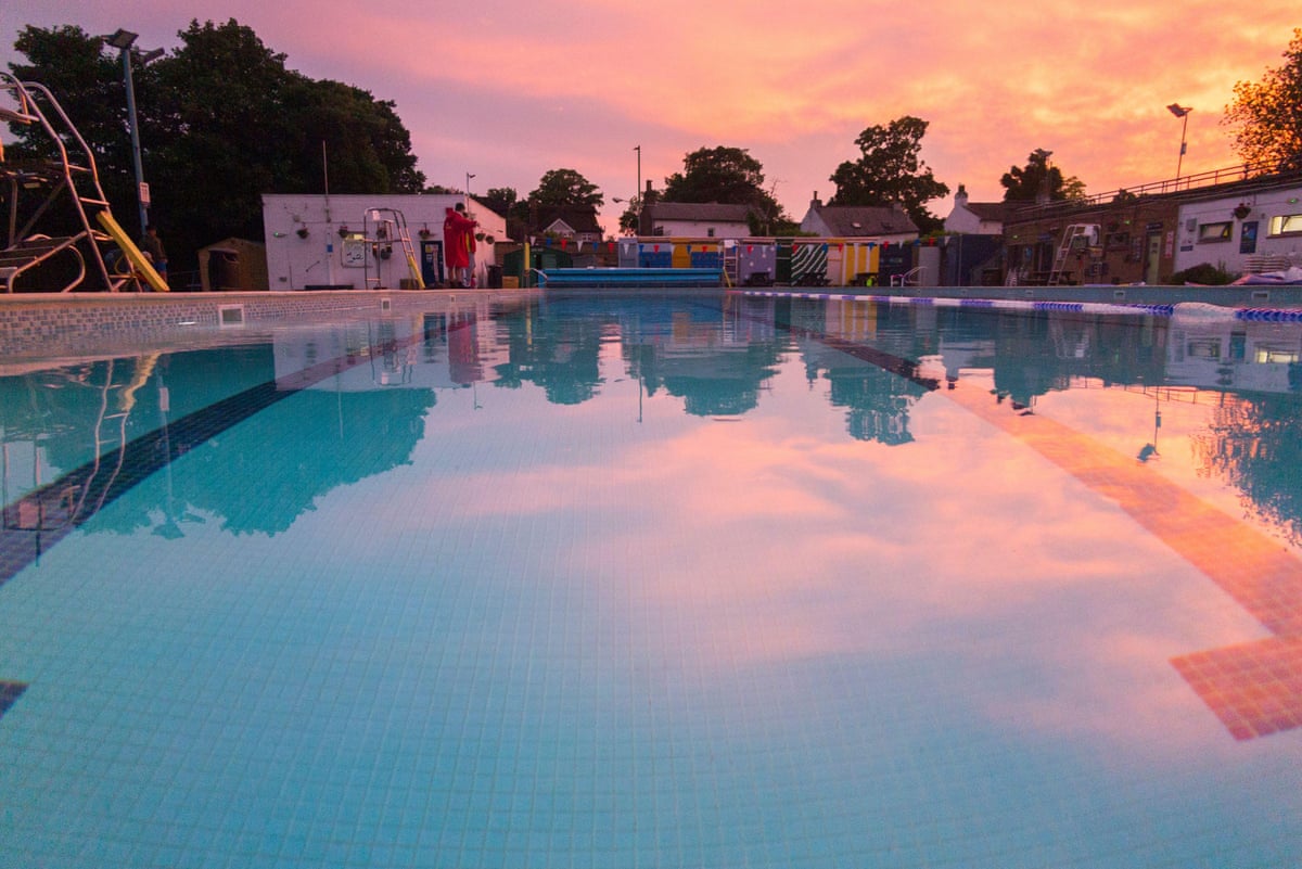 A calm outdoor pool reflecting a pink sunset with trees in the background