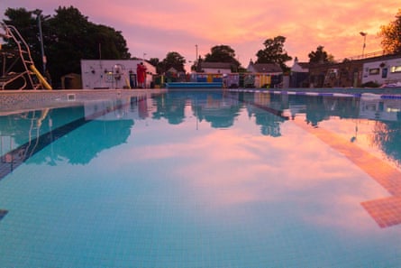 A calm outdoor pool reflecting a pink sunset with trees in the background