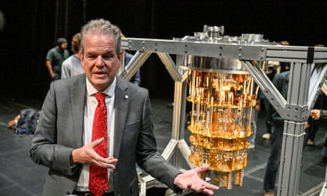 A man in a grey suit and red tie, gesturing with his hands, stands in front of a golden, chandelier-like object