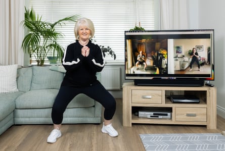A woman with a grey bob doing a workout in a living room, wearing a black tracksuit, with a man and a woman on a split TV screen beside her, doing the same workout