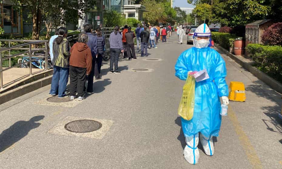 A medical worker in a protective suit walks past people queuing to take a nucleic acid test in a residential area
