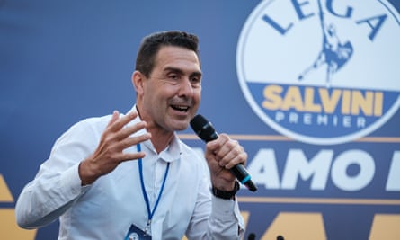 Roberto Vannacci addresses a political rally in Rome; the League symbol and slogans can be seen behind him. He looks animated as he speaks; he is in his mid-50s and has short, neat dark hair, and wears an open-necked pale blue shirt.