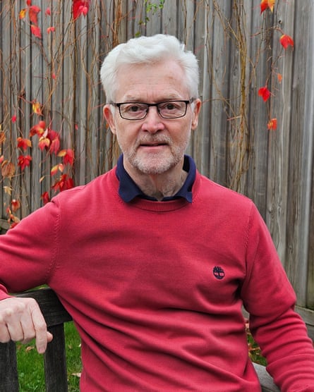 He sits and looks at camera, wearing red jumper