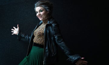 Caitlin Moran poses for a portrait with hands outstretched, wearing a leopard print top, green skirt and black leather jacket, against a black background