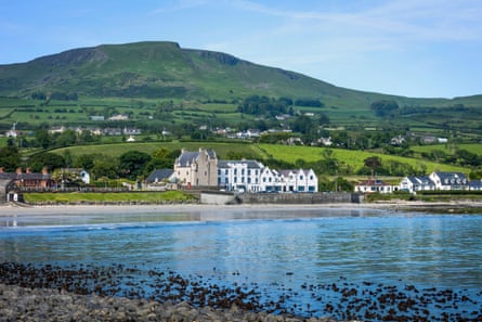 A stone castle next to white hotel buildings with a green hill in the background and water in the foreground