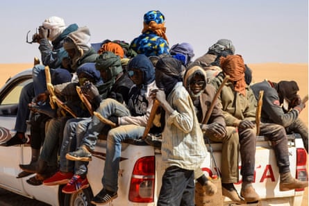 A group of men wearing protective head coverings sit on the back of a truck in the desert
