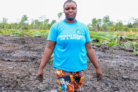 Joy Lebewi, in blue T-shirt, in a muddy area with greenery in the distance.