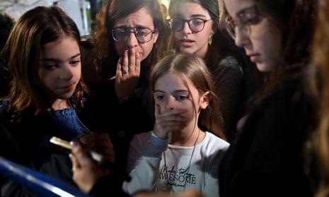 A woman holding a hand to her mouth and four girls look anxiously at a phone screen