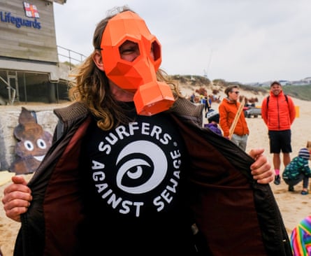 A man on the beach with an orange gas mask on and a sweatshirt saying Surfers Against Sewage