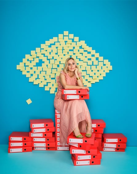 Marina Hyde wearing a floaty peach dress, sitting on top of piles of red box files, with a diamond-shaped collection of blank yellow Post-it notes on a blue wall behind her