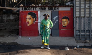 A man wearing a green ANC shawl around his waist and tricorn stands outside a building marked 'Lacoste hair salon'