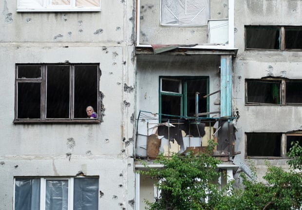 A woman looks out of her apartment window in Sloviansk.  A balcony is destroyed and the walls are covered with dents in the masonry.