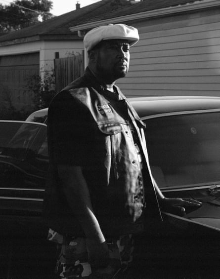 Black and white portrait shows a middle-aged black man with white cap standing beside a car.